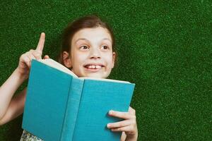 pequeño niña es acostado en artificial césped con un libro foto