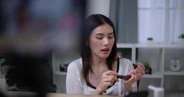 Handheld shot, Beautiful asian woman blogger is showing how to makeup and use cosmetics. In front of the smartphone to record vlog video live streaming at home. Influencer and cosmetics concepts.