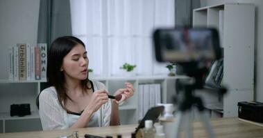 Handheld shot, Beautiful asian woman blogger is showing how to makeup and use cosmetics. In front of the smartphone to record vlog video live streaming at home. Influencer and cosmetics concepts.