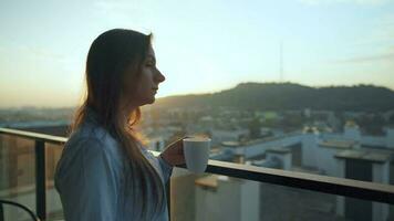 bonito mujer bebidas taza de café o té, acecho un hermosa urbano ver y disfrutar relajarse respiración Fresco congelación aire en balcón a amanecer video