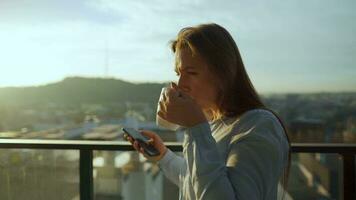 mooi vrouw drankjes kop van koffie of thee, aan het kijken een mooi stedelijk visie en genieten kom tot rust ademen vers bevriezing lucht Aan balkon Bij dageraad video