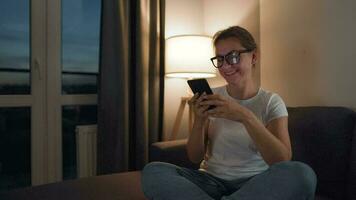 Frau mit Brille Sitzung auf das Sofa im ein gemütlich Zimmer und mit Smartphone zum Surfen Internet im das Abend. sie ist sehr heiter und glücklich. Entspannung und Lebensstil Technologie video