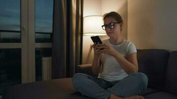 mulher com óculos sentado em a sofá dentro uma acolhedor quarto e usando Smartphone para surfar Internet dentro a tarde. ela é muito alegre e feliz. relaxamento e estilo de vida tecnologia video