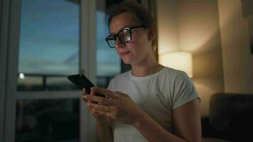 mujer con lentes sentado en el sofá en un acogedor habitación y utilizando teléfono inteligente para surf Internet en el noche. ella es muy alegre y contento. relajación y estilo de vida tecnología video