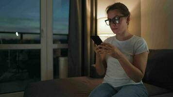 mujer con lentes sentado en el sofá en un acogedor habitación y utilizando teléfono inteligente para surf Internet en el noche. ella es muy alegre y contento. relajación y estilo de vida tecnología video