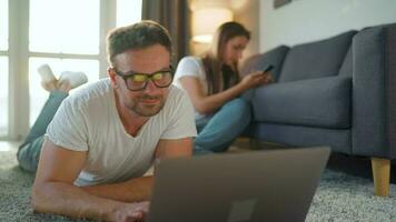 Couple spends time together in a cozy room, man lies on a carpet and uses a laptop, woman with headphones listens to music. Concept of remote work. Home Office video