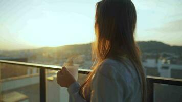 ziemlich Frau Getränke Tasse von Kaffee oder Tee, Aufpassen ein schön städtisch Aussicht und genießen entspannen Atmung frisch Einfrieren Luft auf Balkon beim Dämmerung video