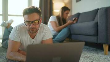 Couple spends time together in a cozy room, man lies on a carpet and uses a laptop, woman with headphones listens to music. Concept of remote work. Home Office video