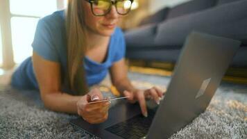 Frau mit Brille ist Lügen auf das Fußboden und macht ein online Kauf mit ein Anerkennung Karte und Laptop. online Einkaufen video
