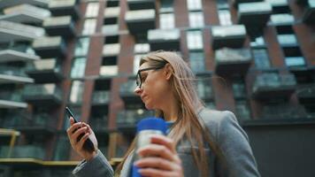 Formally dressed woman with glasses walks down the street in a business district with coffee in hand and uses a smartphone video