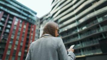 Formally dressed woman with glasses walks down the street in a business district with coffee in hand and uses a smartphone video