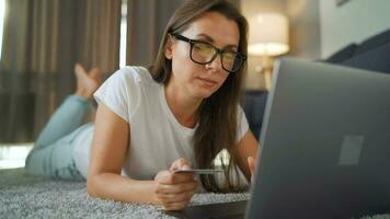 mujer con lentes es acostado en el alfombra y hace un en línea compra utilizando un crédito tarjeta y ordenador portátil. en línea compras, estilo de vida tecnología video