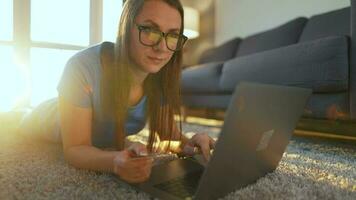 Frau mit Brille ist Lügen auf das Fußboden und macht ein online Kauf mit ein Anerkennung Karte und Laptop. online Einkaufen video