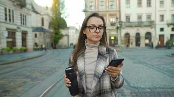 mujer en un Saco y lentes con un termo taza en su mano negociaciones en un teléfono inteligente mientras caminando en el ciudad cuadrado. antiguo europeo arquitectura alrededor. comunicación concepto. video