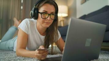 mujer con lentes es acostado en el alfombra y hace un en línea compra utilizando un crédito tarjeta y ordenador portátil. en línea compras, estilo de vida tecnología video