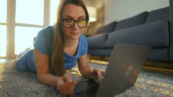 mulher com óculos é deitado em a chão e faz a conectados compra usando uma crédito cartão e computador portátil. conectados compras video