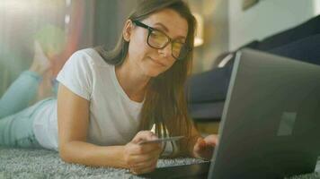 femme avec des lunettes est mensonge sur le tapis et fait du un en ligne achat en utilisant une crédit carte et portable. en ligne achats, mode de vie La technologie video