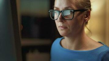 mujer en lentes mirando en el monitor y surf Internet a noche. el monitor pantalla es reflejado en el lentes. trabajo a noche. hogar oficina. remoto trabajo video