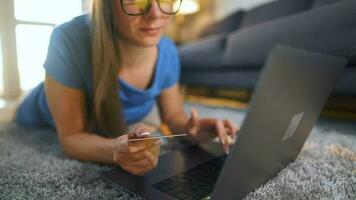 Frau mit Brille ist Lügen auf das Fußboden und macht ein online Kauf mit ein Anerkennung Karte und Laptop. online Einkaufen video