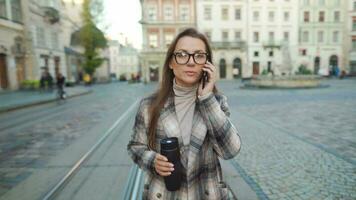 mujer en un Saco y lentes con un termo taza en su mano negociaciones en un teléfono inteligente mientras caminando en el ciudad cuadrado. antiguo europeo arquitectura alrededor. comunicación concepto. video
