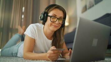 Woman with glasses is lying on the carpet and makes an online purchase using a credit card and laptop. Online shopping, lifestyle technology video