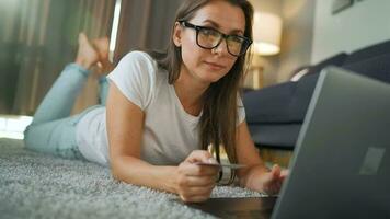 mujer con lentes es acostado en el alfombra y hace un en línea compra utilizando un crédito tarjeta y ordenador portátil. en línea compras, estilo de vida tecnología video