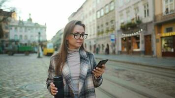 Woman in a coat and glasses with a thermos cup in her hand talks on a smartphone while walking in the city square. Old European architecture around. Communication concept. video