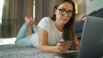 Woman with glasses is lying on the carpet and makes an online purchase using a credit card and laptop. Online shopping, lifestyle technology video