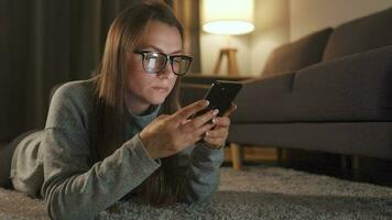 Frau mit Brille ist Lügen auf das Fußboden und mit Smartphone im das Abend video