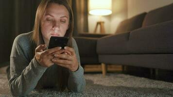 Frau ist Lügen auf das Fußboden und mit Smartphone im das Abend video