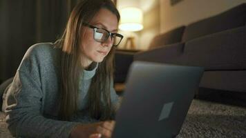 Frau mit Brille ist Lügen auf das Teppich und Arbeiten auf ein Laptop im das Abend. Konzept von Fernbedienung Arbeit video