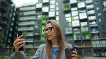 Formally dressed woman with glasses walks down the street in a business district with coffee in hand and uses a smartphone video