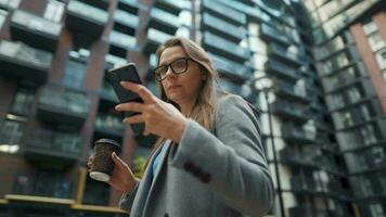 Formally dressed woman with glasses walks down the street in a business district with coffee in hand and uses a smartphone video