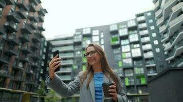Formally dressed woman with glasses walks down the street in a business district with coffee in hand and uses a smartphone video