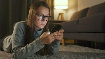 Woman with glasses is lying on the floor and using smartphone in the evening video