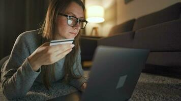 mujer con lentes es acostado en el piso y hace un en línea compra utilizando un crédito tarjeta y ordenador portátil en el noche video