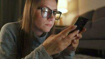 Woman with glasses is lying on the floor and using smartphone in the evening video