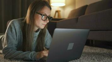 Woman with glasses is lying on the carpet and working on a laptop in the evening. Concept of remote work video