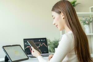 Investor analyzing exchange market, asian young business woman trader on graphic charts on laptop computer, hand in using smartphone for trading stock, looking at screen with diagrams at home office. photo