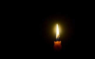 una sola llama de vela encendida o luz que brilla en una vela naranja sobre fondo negro u oscuro en la mesa de la iglesia para Navidad, funeral o servicio conmemorativo con espacio para copiar foto