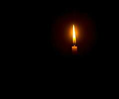 A single burning candle flame or light glowing on an orange candle on black or dark background on table in church for Christmas, funeral or memorial service with copy space photo