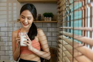 Healthy asian woman drinking a glass of dairy milk for while wearing sportswear for nutrition and protein supplement at the window for pregnancy and maternity benefit concept photo