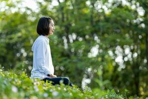 asiático mujer es relajantemente practicando meditación yoga en bosque lleno de salvaje flor en verano a alcanzar felicidad desde interior paz sabiduría con Mañana ligero para sano mente y alma concepto foto