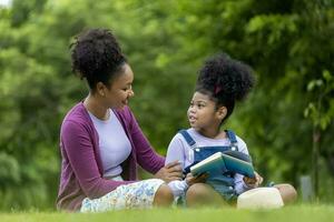 africano americano madre es enseñando su joven hija a leer mientras teniendo un verano picnic en el público parque para educación y felicidad foto