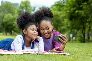 africano americano madre y joven hija son acostado abajo y tomando selfie foto durante verano picnic en el público parque para fin de semana ocio y felicidad concepto