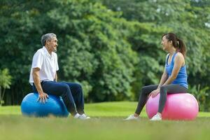 Senior asian man and his daughter is sitting on the yoga ball in the public park while having leisure conversation together for elder longevity exercise and outdoor workout usage photo