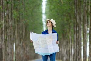 asiático turista mujer es mirando a el mapa para dirección mientras teniendo vacaciones a el nacional parque mientras caminando en el la carretera con columna de árbol para viaje y aventuras concepto foto