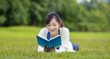 Asian woman is lying down in the grass lawn inside the public park holding book in her hand during summer for reading and education concept photo