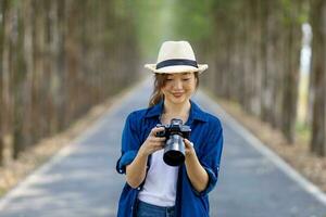 Asian tourist woman is taking photo using professional camera while having vacation at the national park while walking on the road with column of tree for travel and photography concept