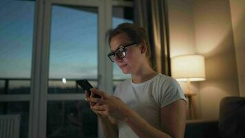 Frau mit Brille Sitzung auf das Sofa im ein gemütlich Zimmer und mit Smartphone zum Surfen Internet im das Abend. sie ist sehr heiter und glücklich. Entspannung und Lebensstil Technologie video